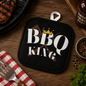 Black pot holder with 'BBQ King' text on a wooden table with grill items.